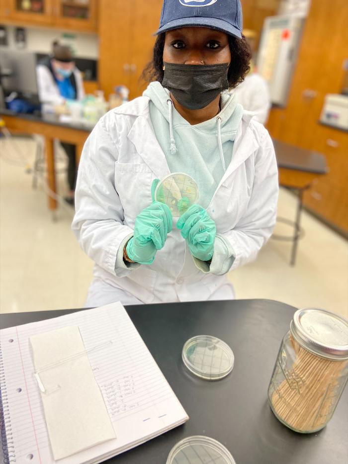 在生物实验室里，一名学生穿着个人防护装备，拿着一个培养皿，培养皿上生长着细菌