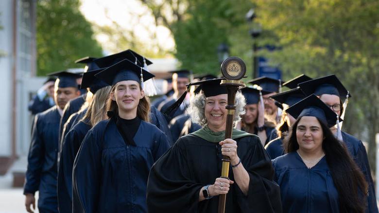 毕业生们头戴帽子，身穿长袍，排成两排在人行道上走着，由身穿黑袍的教员领着，手里拿着一根手杖和一个吊坠.